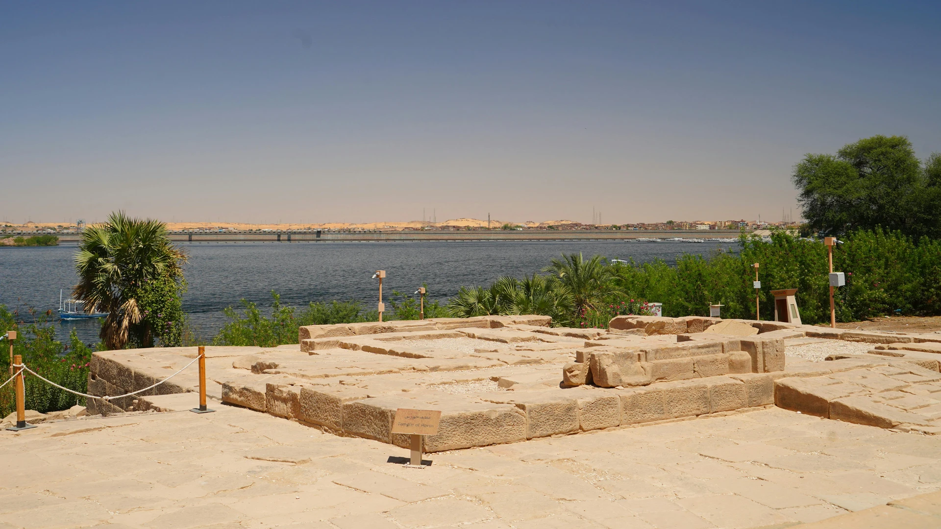 Aswan, Egypt - View of ancient ruins near the nile river surrounded by greenery and distant structures under a clear blue sky.
